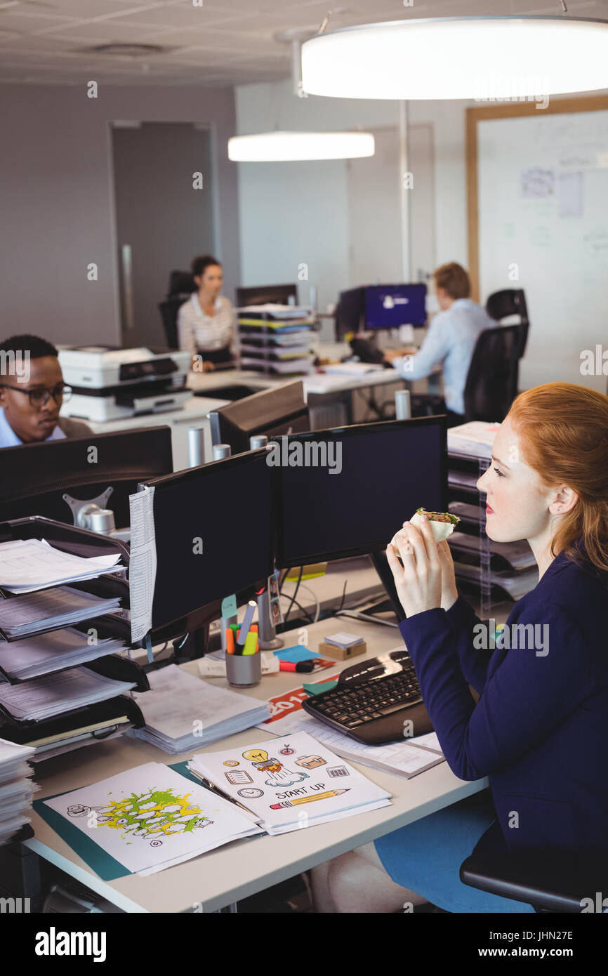 Imprenditrice di mangiare uno spuntino al banco mentre i colleghi che lavorano in ufficio creativo Foto Stock