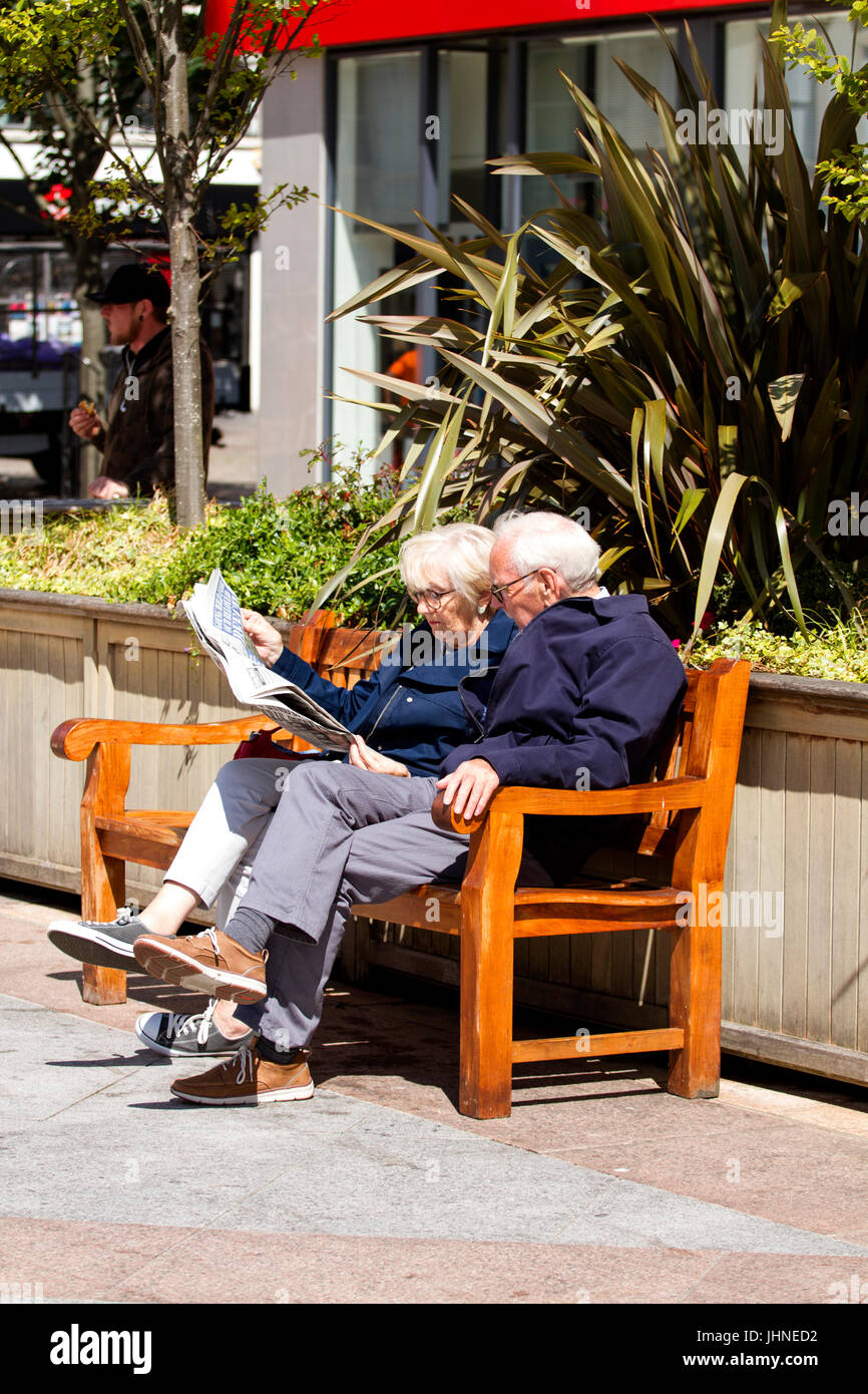 Una coppia di anziani seduti su un sedile insieme leggendo un giornale presso la piazza della città di Dundee, Regno Unito Foto Stock