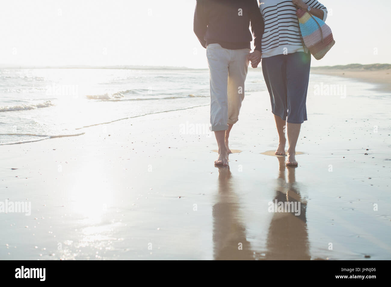 Affettuoso a piedi nudi coppia matura a piedi, tenendo le mani nella soleggiata ocean beach surf Foto Stock