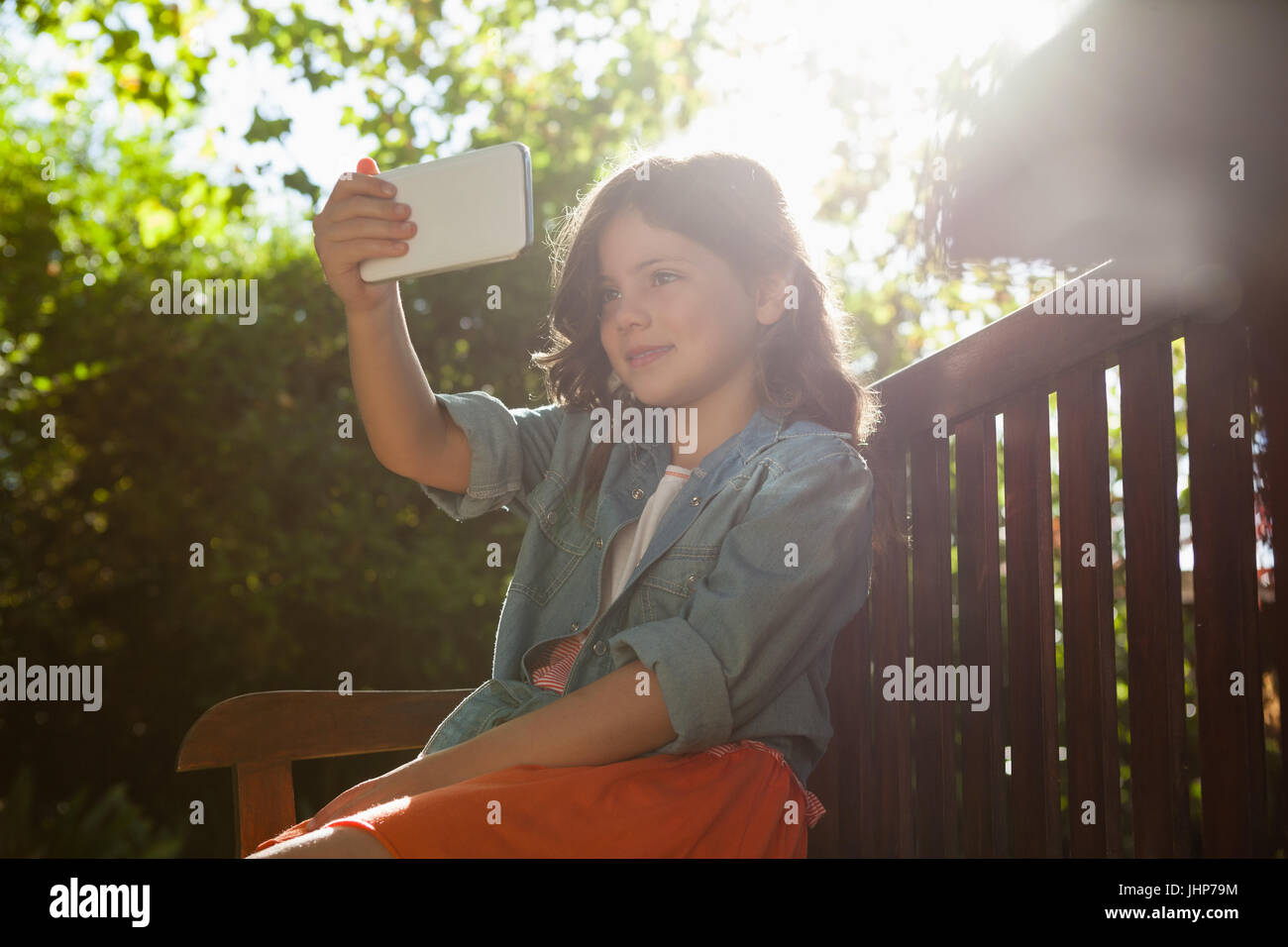 Ragazza tenendo selfie dal telefono cellulare mentre è seduto sulla panca di legno a backyard durante la giornata di sole Foto Stock
