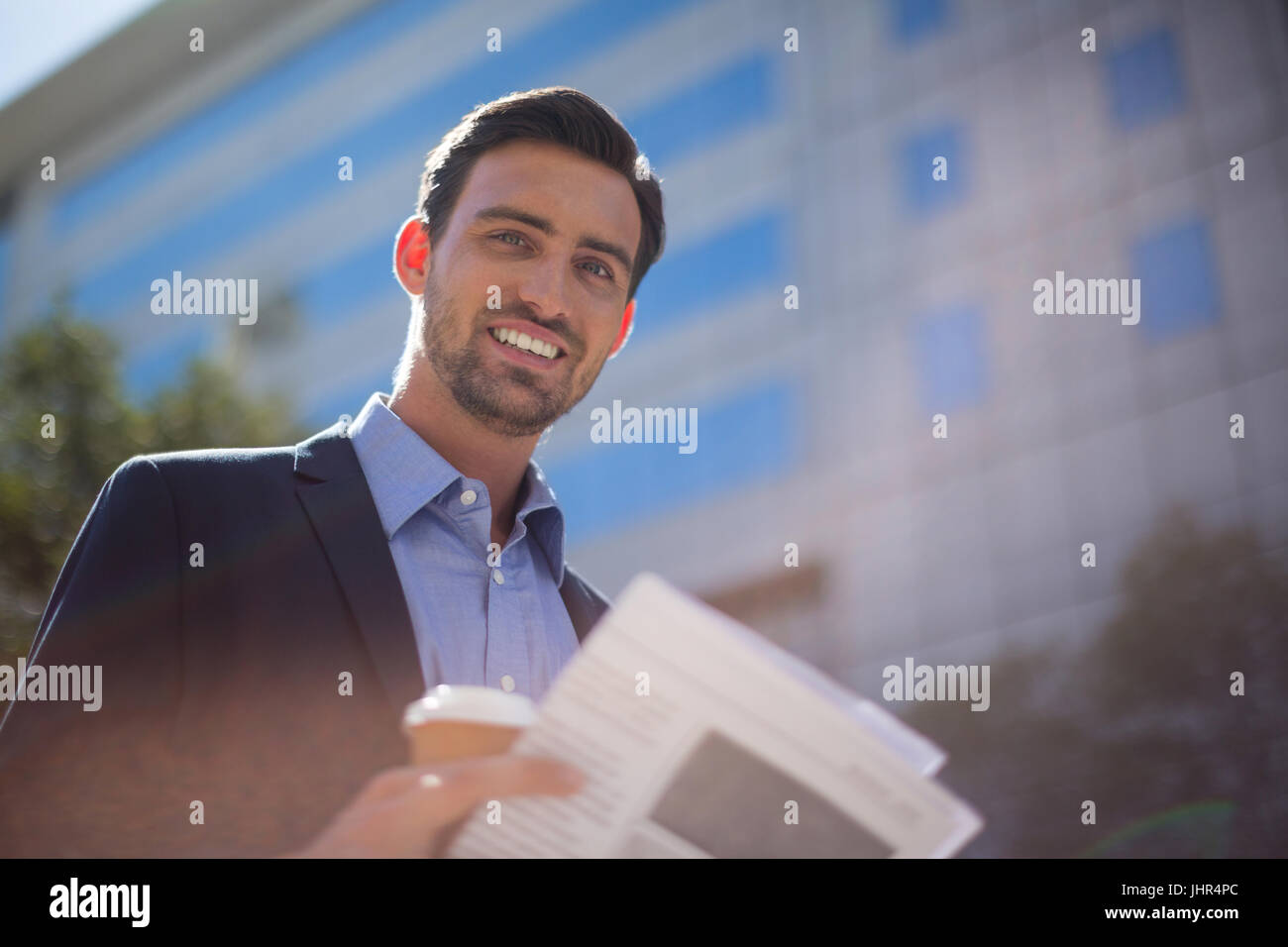 Ritratto di imprenditore sorridente azienda giornale e la tazza di caffè vicino edificio per uffici Foto Stock