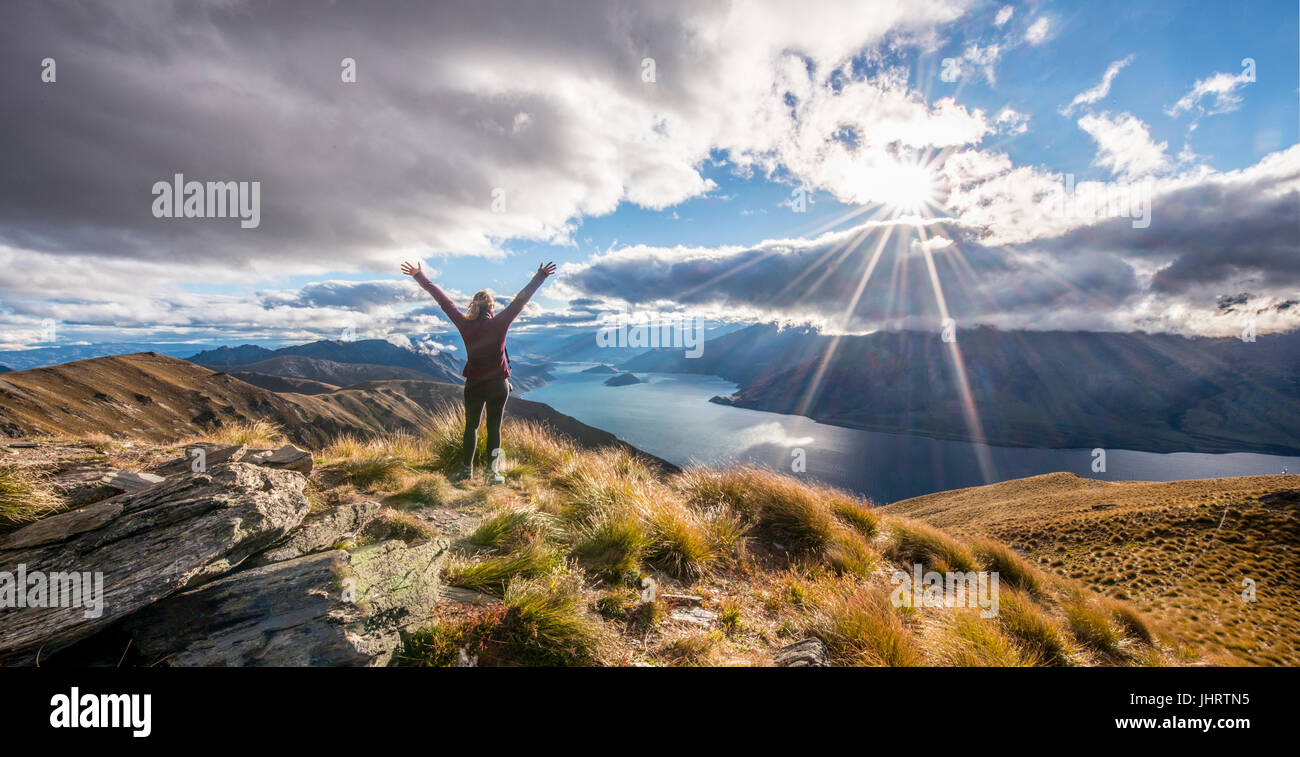 Escursionista bracci di estensione nell'aria, il sole splende, il lago Wanaka e paesaggio di montagna, Istmo di picco, Otago, Isola del Sud Foto Stock