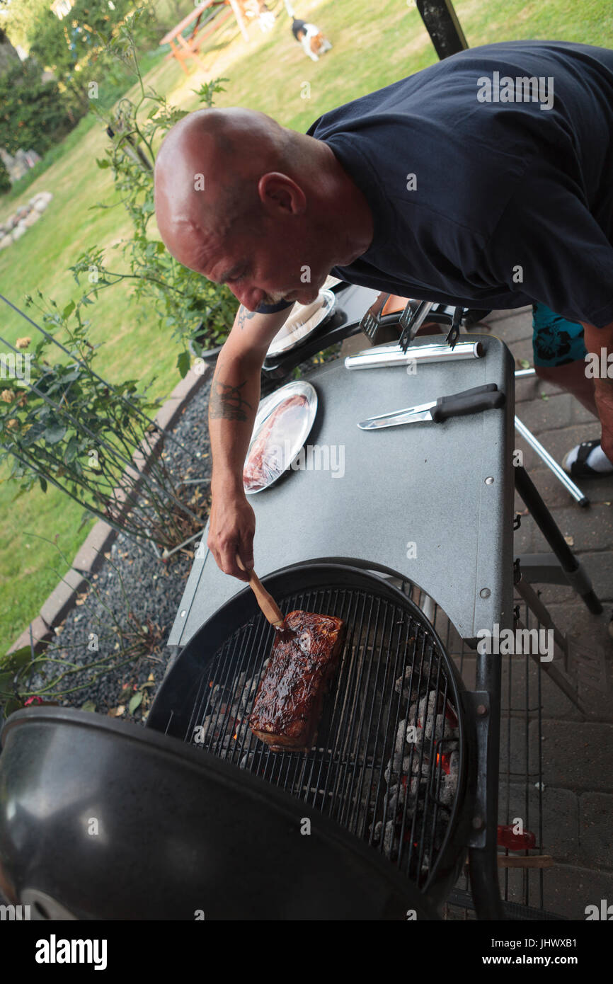 L'uomo senza capelli inumidendo di carne su un carbone di legna per barbecue cucinare sopra il calore indiretto sulle braci, cane in background in giardino Foto Stock
