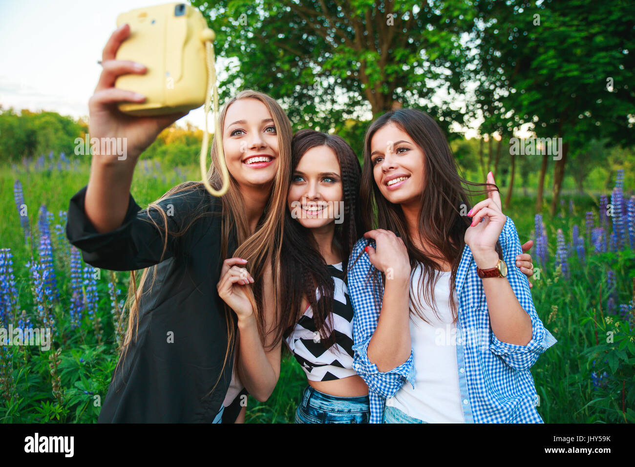 Tre ragazze hipsters bionda e bruna tenendo autoritratto sulla fotocamera polaroid e sorridente all'aperto. Le ragazze per divertirsi insieme nel parco. Foto Stock