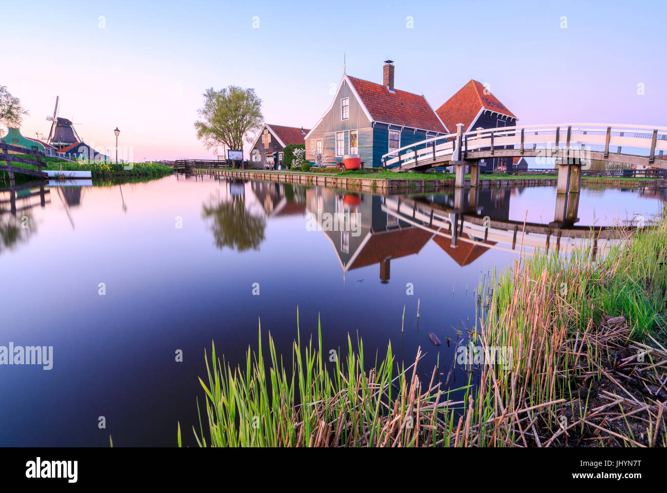 Case in legno e il mulino a vento si riflette nel blu fiume Zaan al tramonto, Zaanse Schans, North Holland, Paesi Bassi, Europa Foto Stock