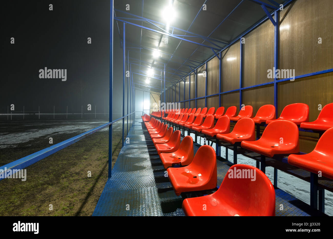 Vuoto tribune con righe rosse umido sedili in plastica su un campo di calcio con la neve e pozzanghere. (A NOTTE) Foto Stock