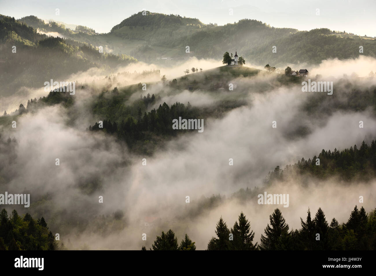 Nebbia di rotolamento a sunrise nel Skofjelosko Hribovje colline con la chiesa di San Tommaso sulla collina vicino a Skofja Loka Slovenia Foto Stock