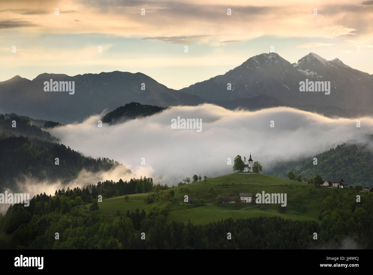 Nebbia di rotolamento a sunrise nel Skofjelosko colline con la chiesa di San Tommaso con montagne di Kamnik Savinja Alpi vicino a Ljubljana Slovenia Foto Stock