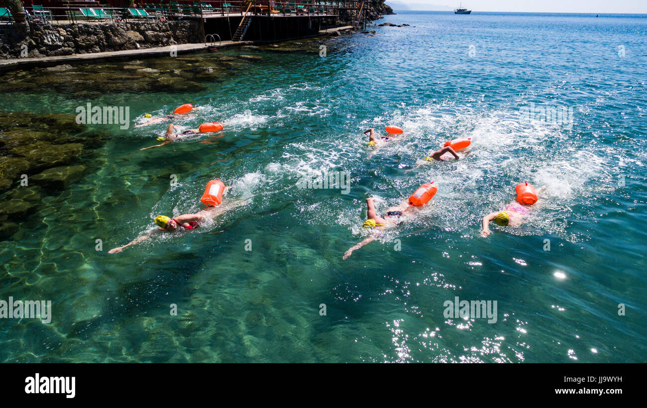 Il gruppo di sette giovani nuotatori - ciascuna con un salvagente - formazione nelle limpide e trasparenti acque del mar Ligure con terrazza e pontoon dietro di loro e una barca in background in una giornata di sole nei paraggi, Cinque Terre, Italia Foto Stock