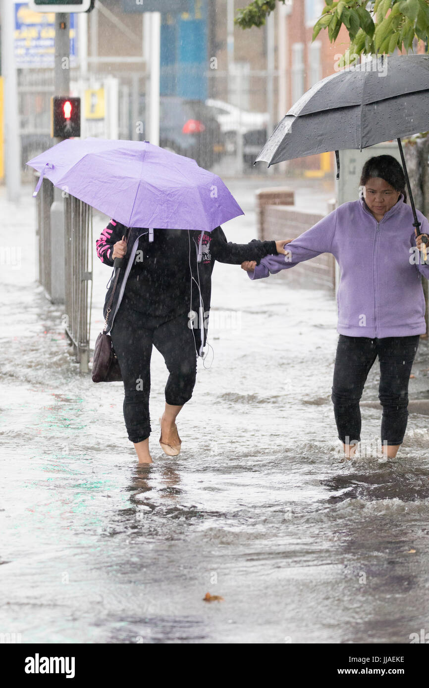 Due donna a piedi attraverso il flash l'acqua di allagamento sul loro modo home dopo forti piogge ha causato allagamenti per la A525 in estate durante il tuono e le tempeste di fulmini che ha colpito la zona, Rhyl, Galles del Nord, Regno Unito Foto Stock