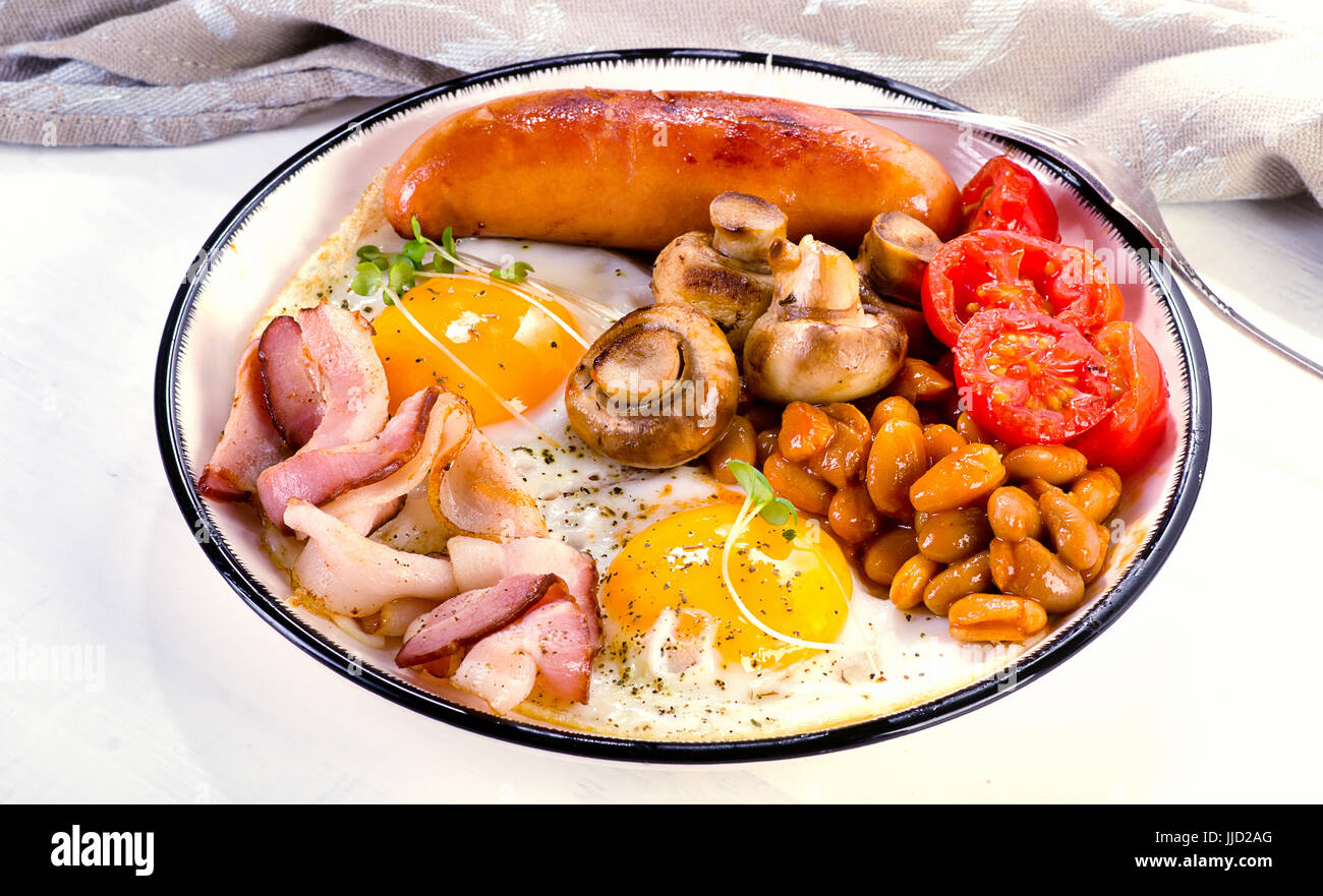 La completa prima colazione inglese con uovo fritto, fagioli, pomodori, funghi e pancetta e salsiccia sulla piastra bianca Foto Stock