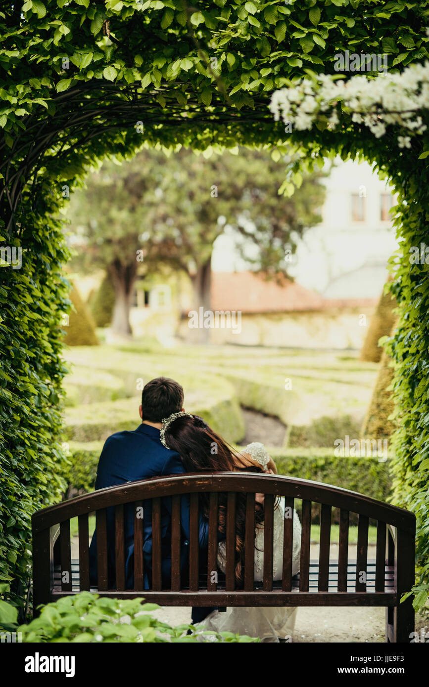 La magnifica vista della sposa giovane di trascorrere del tempo in giardino. Appena sposati sono seduta sul banco sotto il verde blooming arch a Praga. Foto Stock