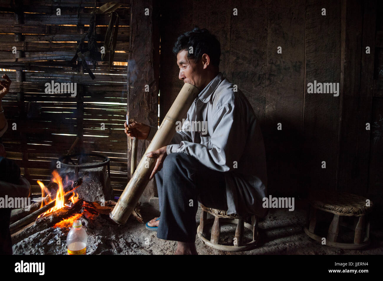 Un uomo fuma una sigaretta attraverso un bambù grande acqua bong da un incendio in una casa di Ban Sop Kha, Laos. Foto Stock