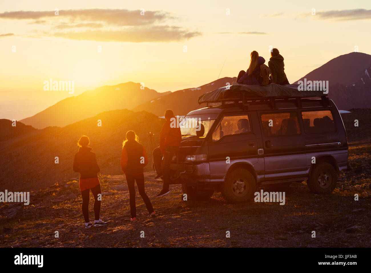 Amicizia e concetto di viaggio con un gruppo di cinque amici seduti e in piedi accanto a auto sulla cima della montagna al tramonto Foto Stock