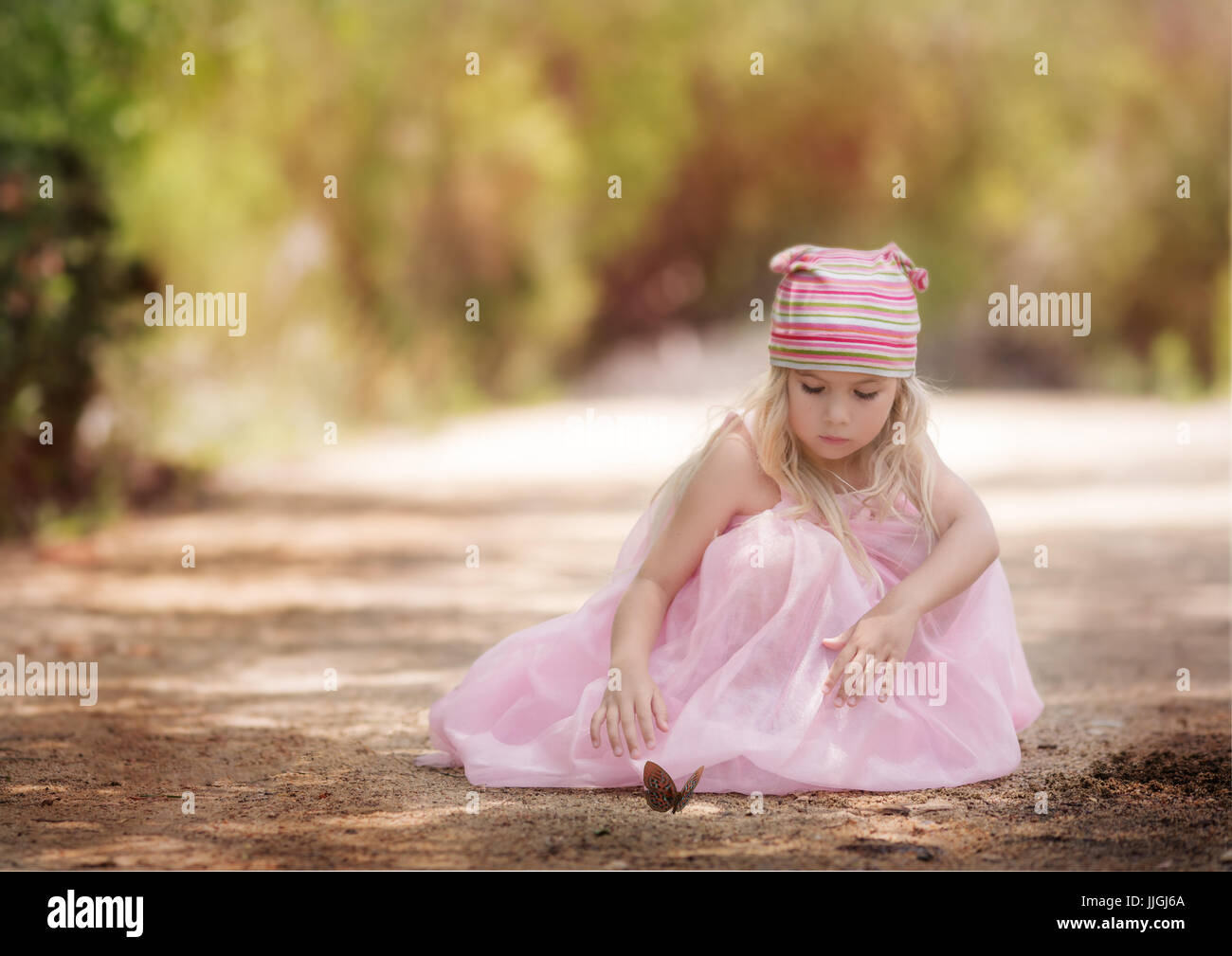 Ragazza in un vestito vintage e cappello di lana seduta su una strada sterrata che guarda una farfalla, California, Stati Uniti Foto Stock
