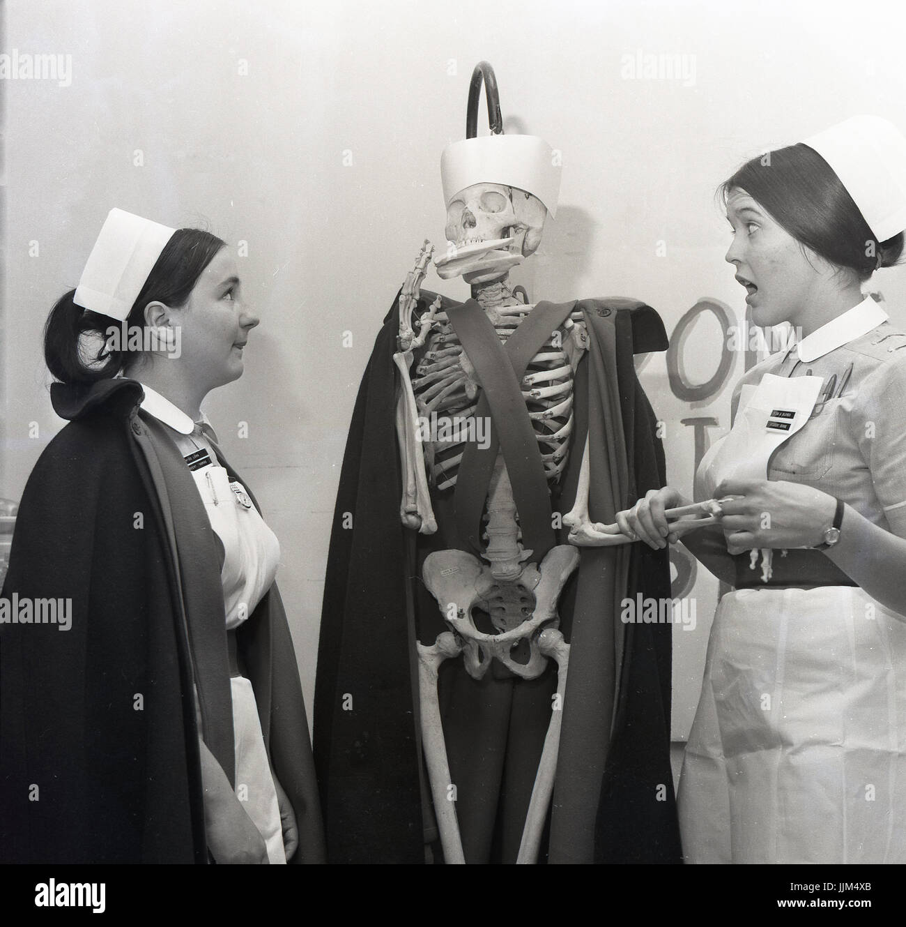 "È ancora vivo?" degli anni sessanta, in Inghilterra, in un ospedale, una femmina di infermiere finge di controllare il polso di una dimensione di vita scheletro modello vestito con un infermiere del promontorio e cappello e con un sandwich in bocca. La foto è stata per un giornale locale per aumentare la consapevolezza circa il basso dei salari per infermieri. Foto Stock