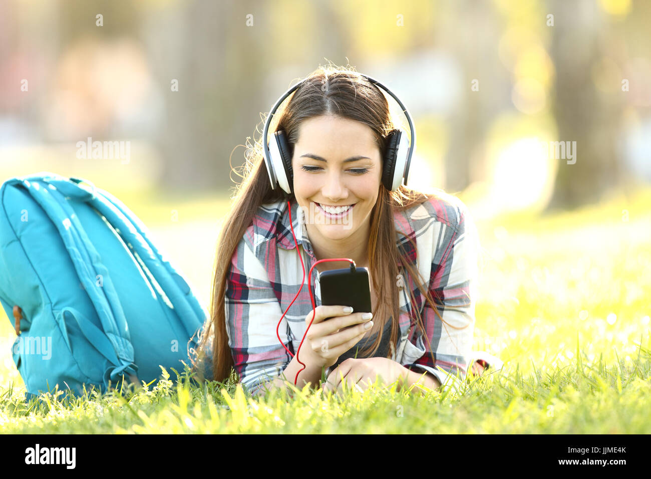 Felice studente ascolto lezioni audio in linea con le cuffie e uno smart phone sdraiati sull'erba in un campus Foto Stock