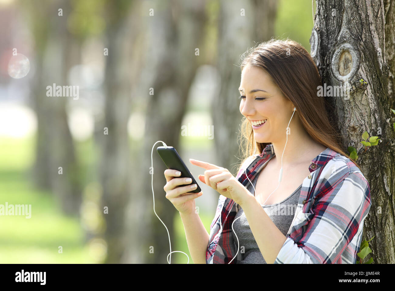 Bella ragazza teen di ascolto e di scelta della musica in uno smart phone appoggiato su di un albero in un parco Foto Stock