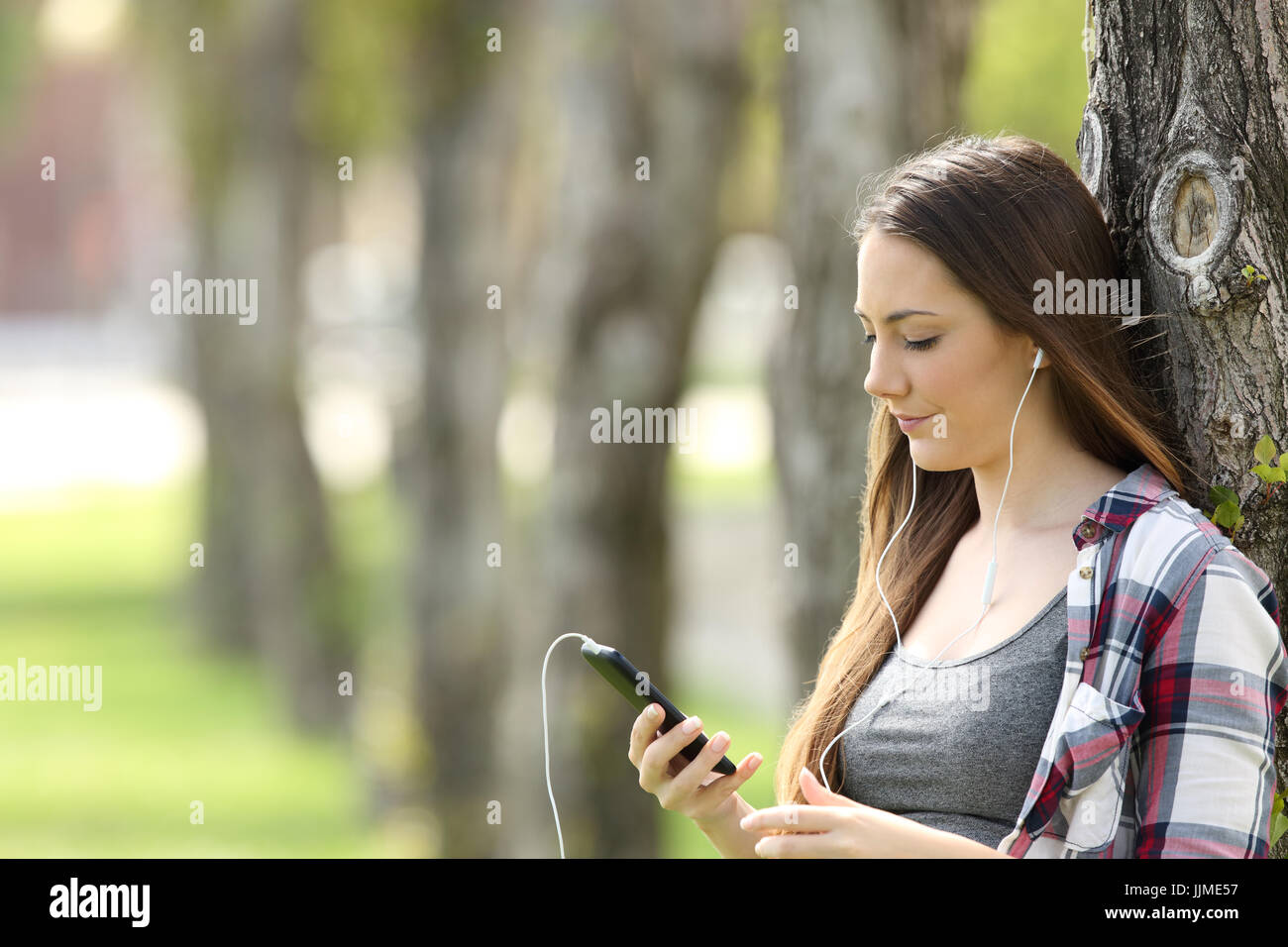 Rilassati ascoltando la ragazza sulla linea di musica e la visione di contenuti multimediali appoggiato su di un albero in un parco Foto Stock