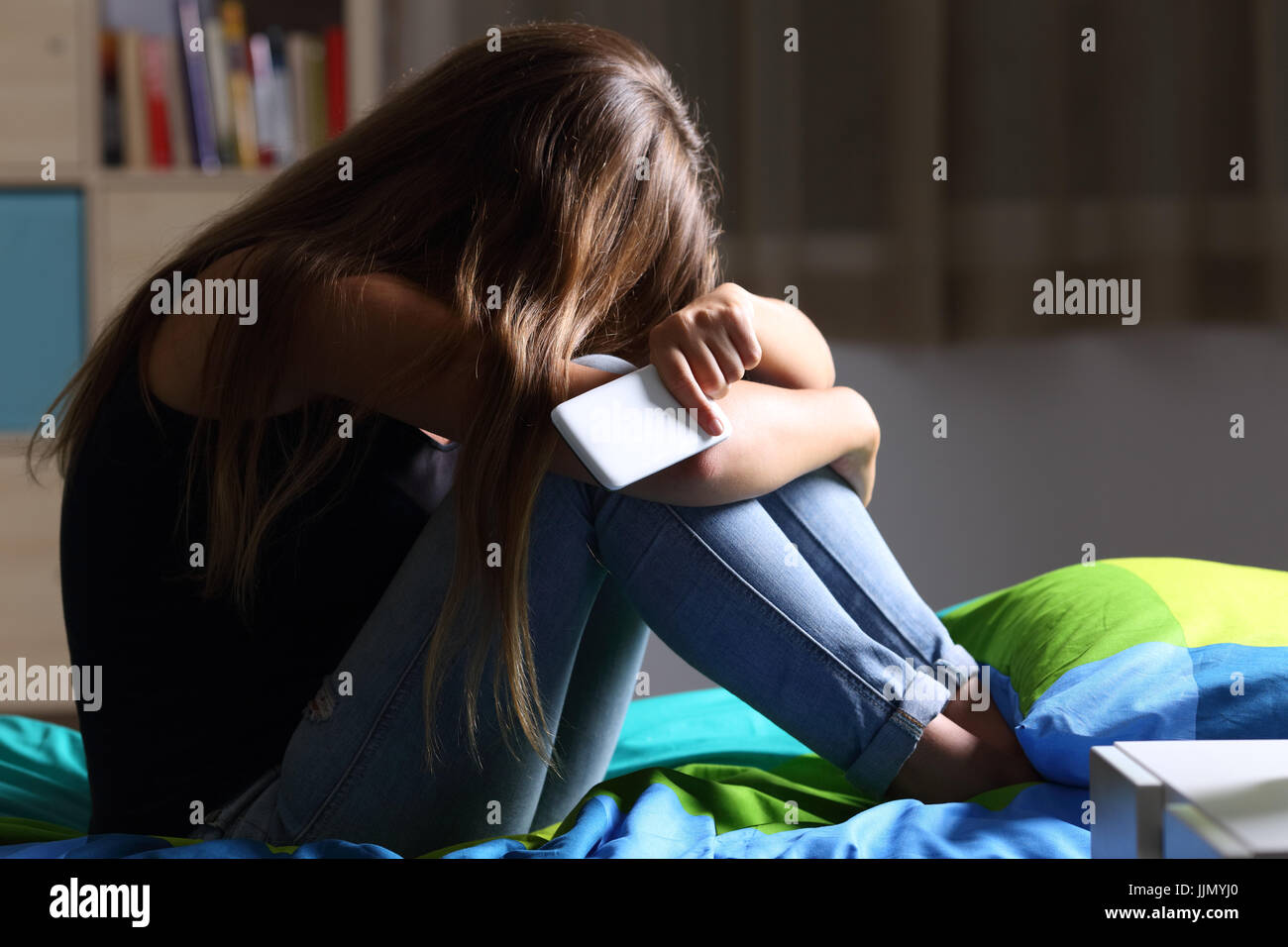 Unico triste teen in possesso di un telefono cellulare piange seduta sul letto nella sua camera da letto con una luce oscura in background Foto Stock