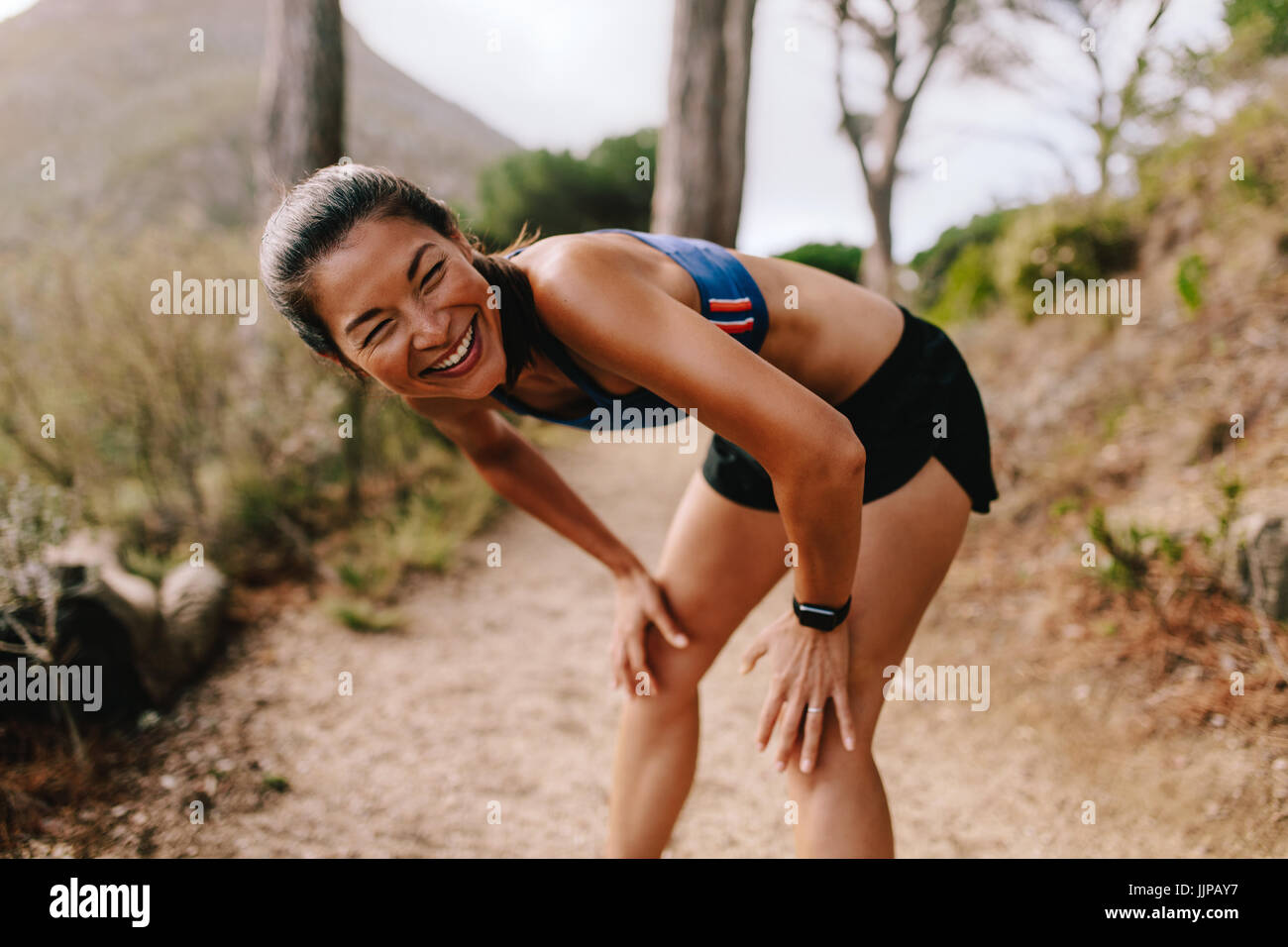 Montare la giovane donna asiatica in piedi sul sentiero di montagna con le sue mani sulle ginocchia e sorridente. Femminile in sportswear prendendo una pausa dopo l'esecuzione di allenamento Foto Stock