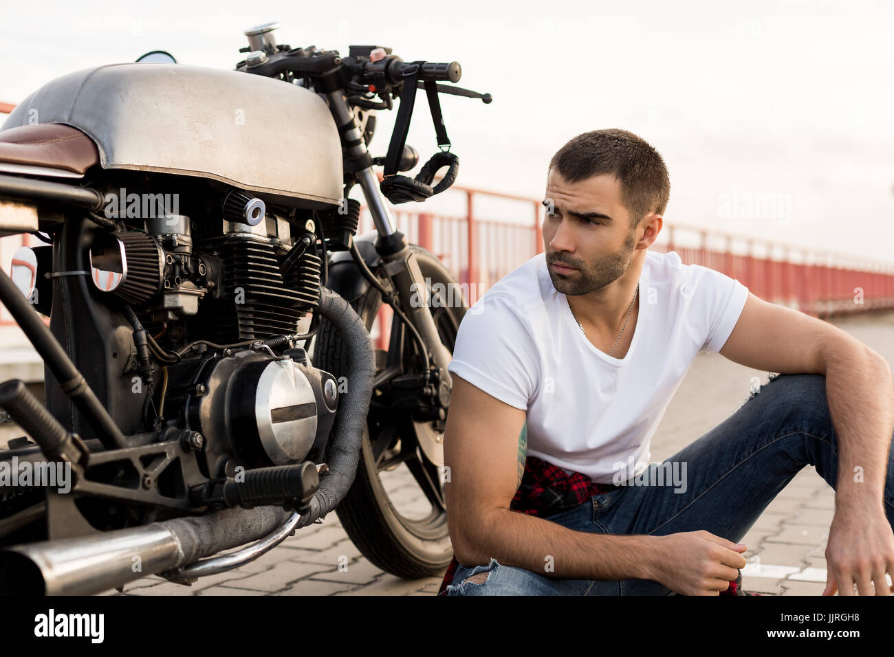 Bello felice cavaliere ragazzo con barba e baffi seduto vicino al suo stile classico biker cafe racer moto e guardare al lato. Moto custom made in vint Foto Stock