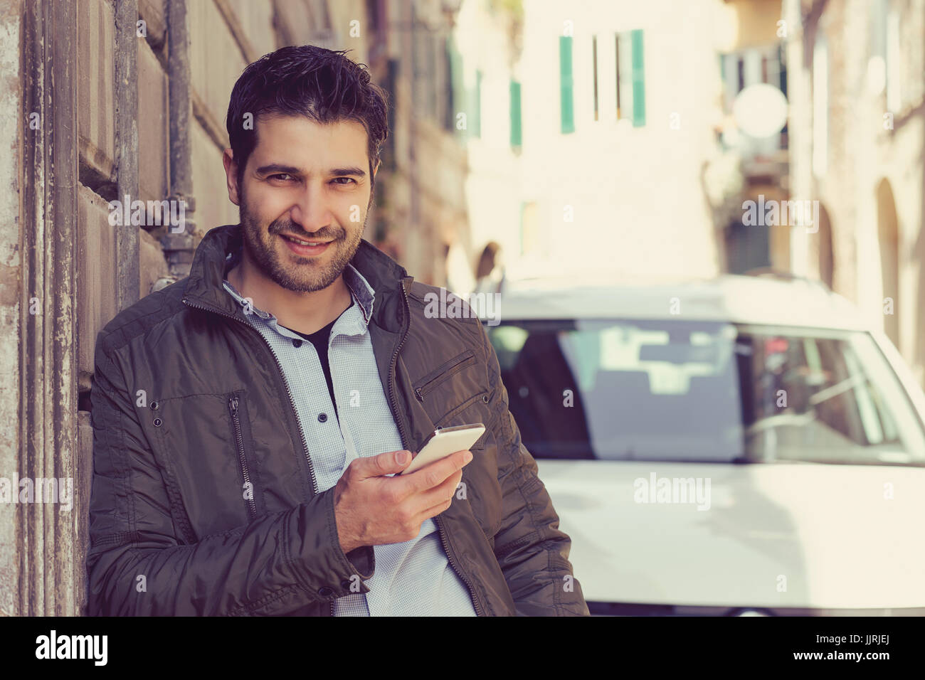 Uomo sorridente in possesso di un telefono cellulare all'aperto in piedi accanto alla sua nuova vettura texting. Foto Stock