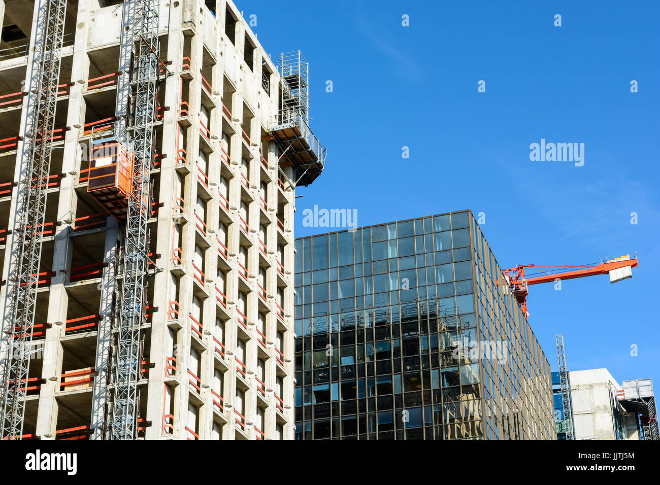 Basso angolo di visione di un edificio di vetro tra due edifici in cemento in fase di costruzione con una torre rossa gru contro il cielo blu. Foto Stock