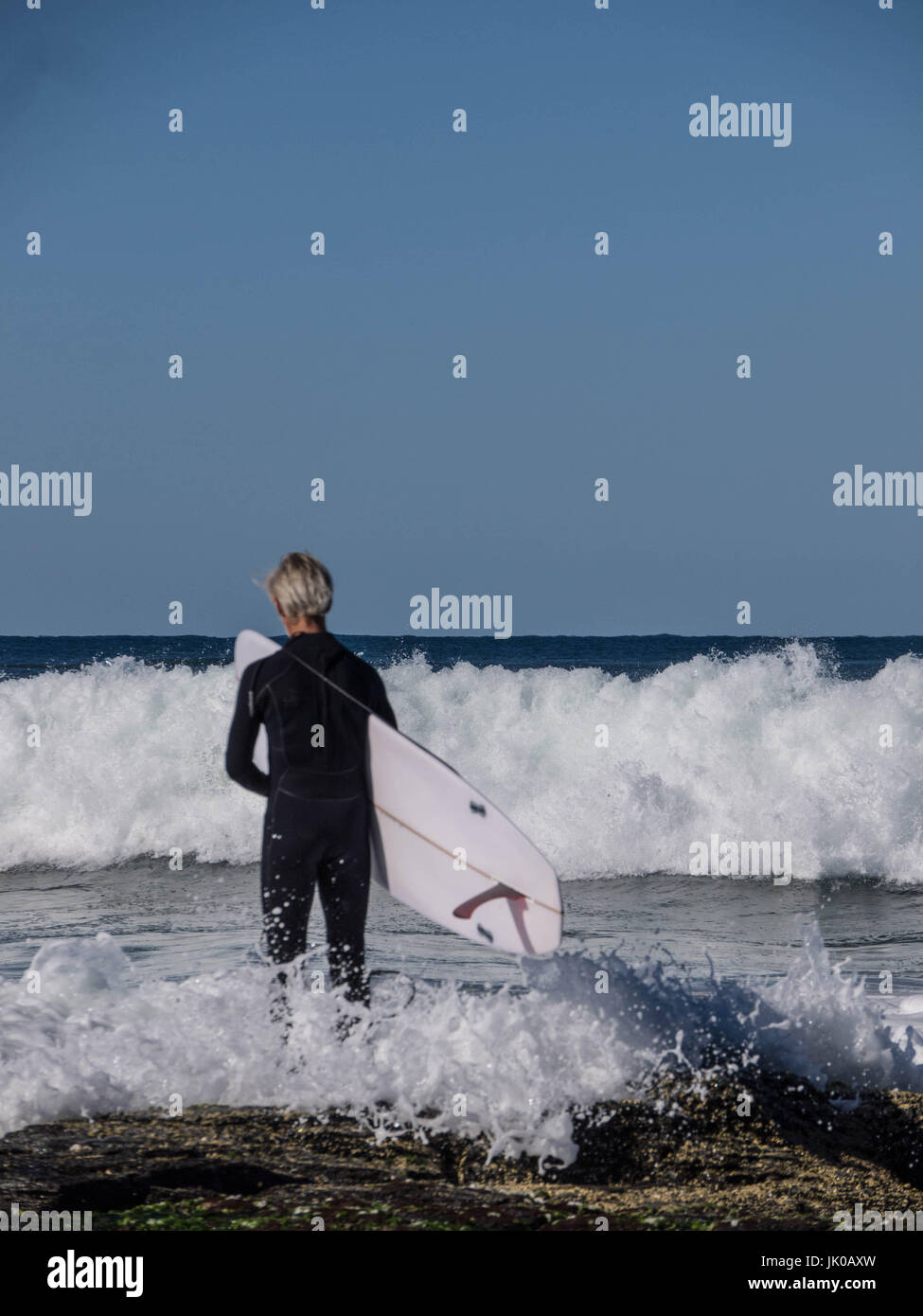 Un vecchio surfista maschio in piedi e in attesa per le Onde Shorebreak tenendo la sua tavola da surf sulle rocce dall'oceano Foto Stock