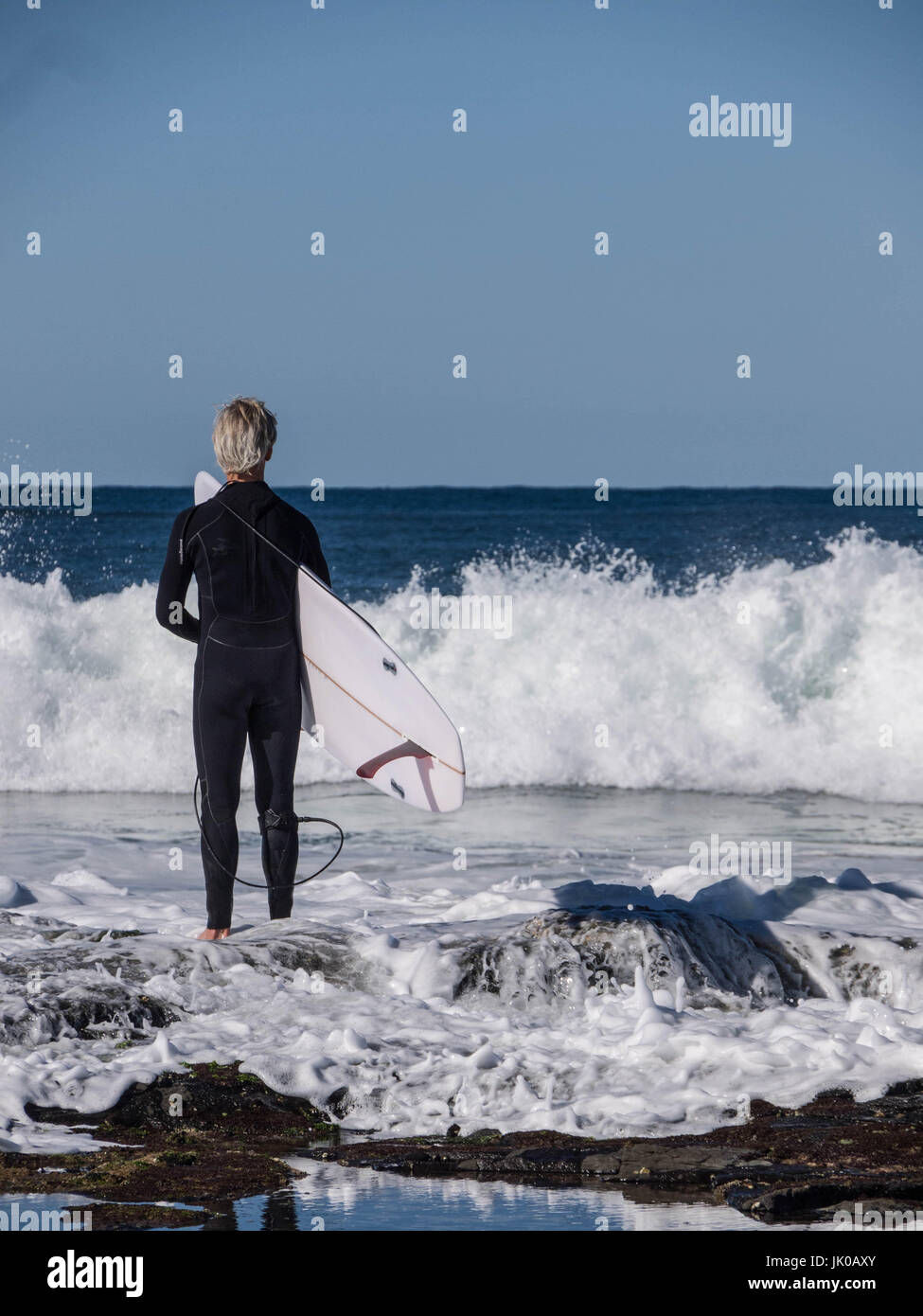 Un vecchio surfista maschio in piedi e in attesa per le Onde Shorebreak tenendo la sua tavola da surf sulle rocce dall'oceano Foto Stock