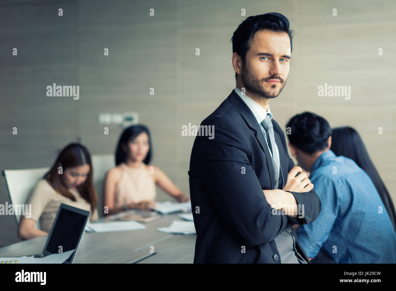Happy business man in piedi con le braccia incrociate in ufficio mentre i colleghi a discutere in background. Foto Stock