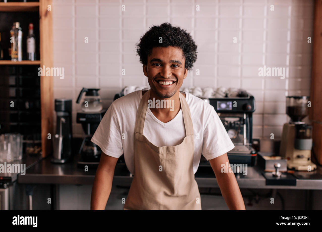 Ritratto di felice giovane barista permanente al cafe contatore. L'uomo africano nel grembiule guardando la fotocamera e sorridente. Foto Stock