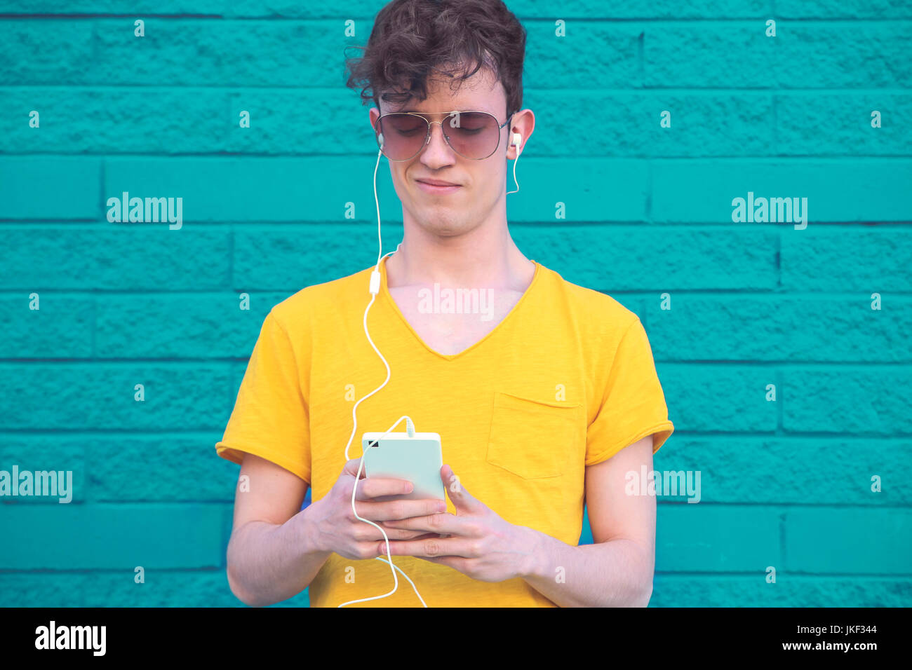 Ritratto di giovane uomo che ascolta musica con lo smartphone e gli auricolari di fronte blu muro di mattoni Foto Stock