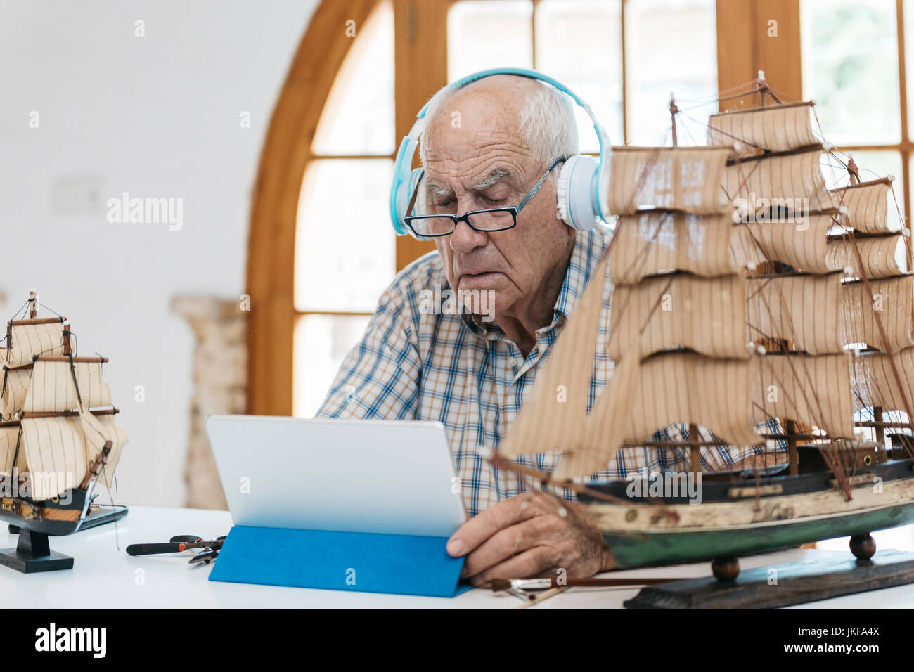 Senior uomo che indossa le cuffie lavorando sul modello di nave sul tavolo con la compressa Foto Stock