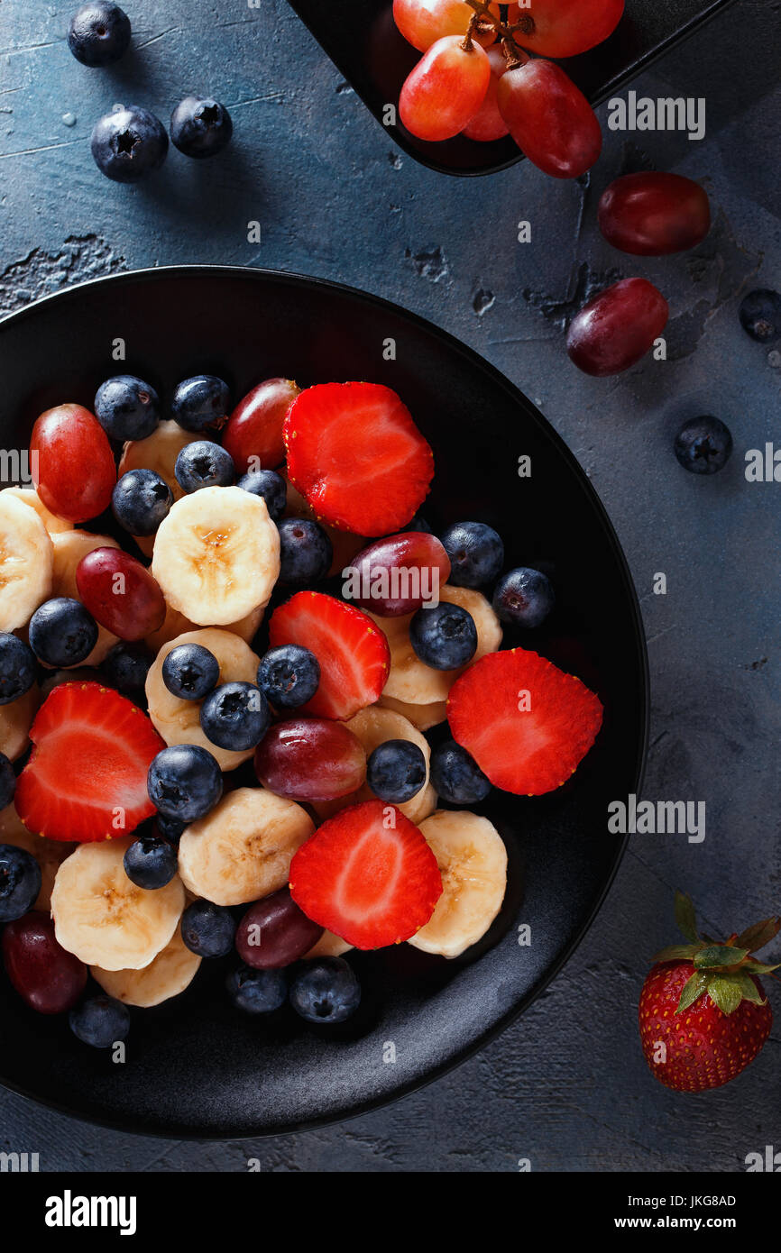 Utile prima colazione di frutti e bacche, vista dall'alto. Fette di banane, fragole, uva e mirtilli in una piastra nera Foto Stock