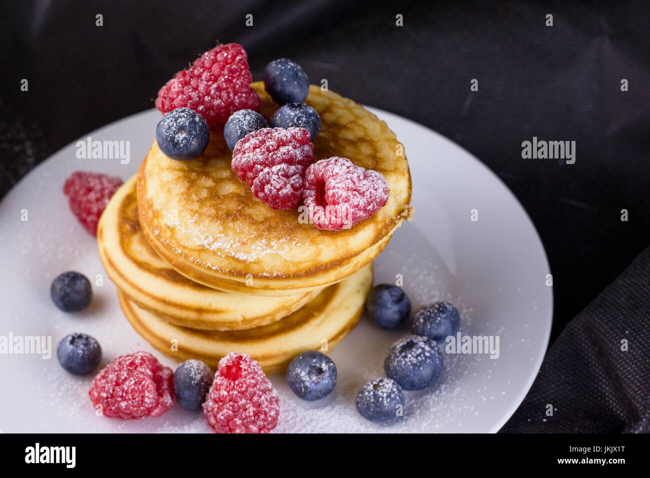 Pila di pancake con lamponi e mirtilli rivestite con zucchero a velo sulla piastra bianca e sfondo nero Foto Stock