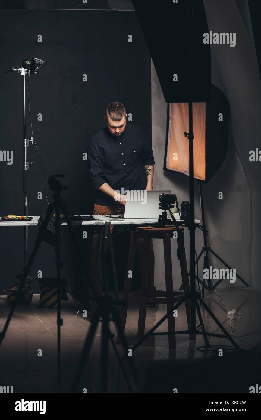 Fotografo lavorando sul computer portatile mentre in piedi dal cibo contro la piastra di sfondo allo studio Foto Stock