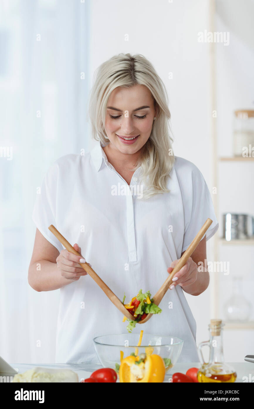 Sorridente giovane donna tossing insalata fresca sul banco di cucina a casa Foto Stock