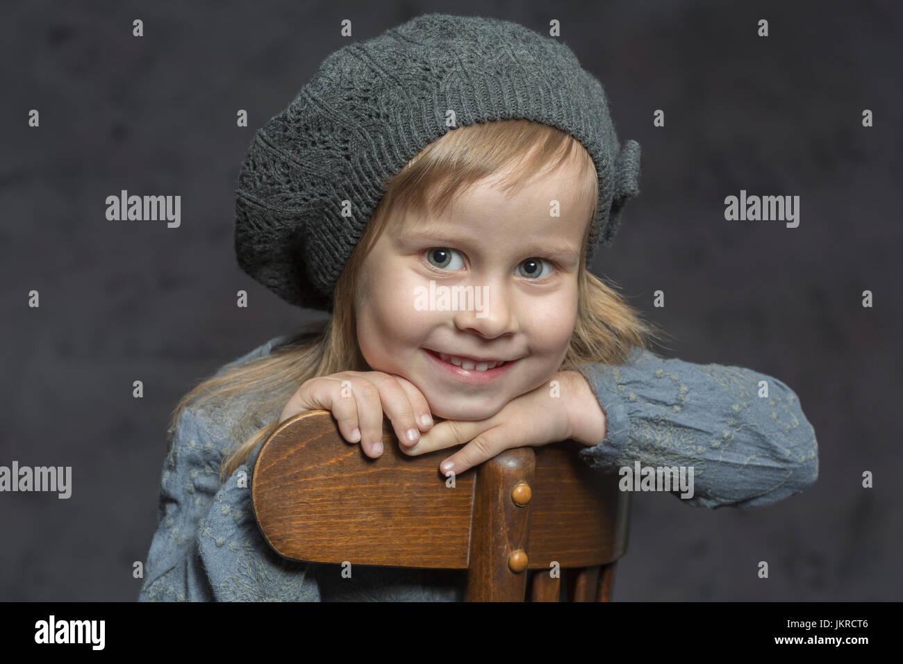 Ritratto di ragazza sorridente indossando knit hat seduto sulla sedia di legno contro uno sfondo grigio Foto Stock