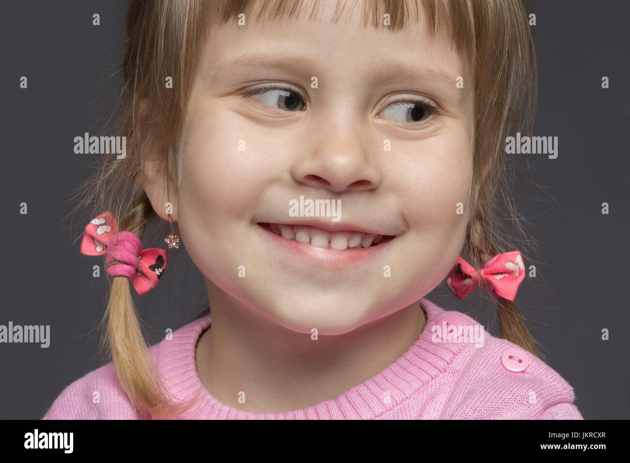 Close-up di carino sorridente bambina indossa un maglione rosa che guarda lontano contro lo sfondo grigio Foto Stock