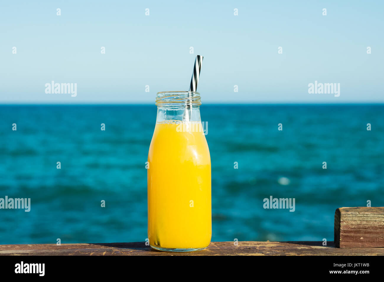 Bottiglia di vetro con succhi di agrumi arancio mandarino con paglia sulla rampa di legno, turchese del mare e cielo blu in background, impostazione naturale, vacati Foto Stock