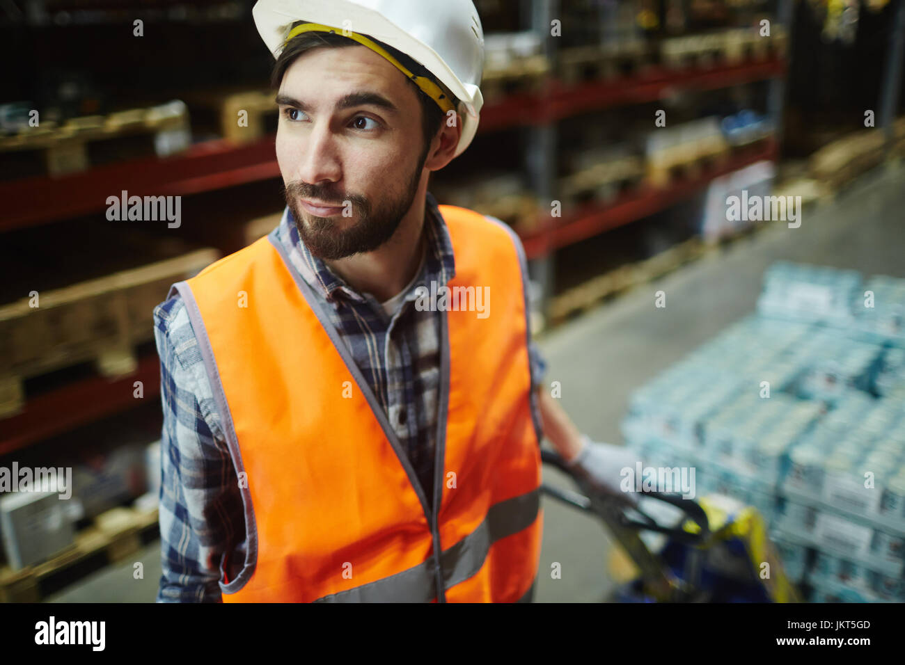 Ritratto di barbuto di lavoro del caricatore in magazzino, tirando il carrello in movimento con prodotti venduti al dettaglio nel corridoio tra gli scaffali alti Foto Stock