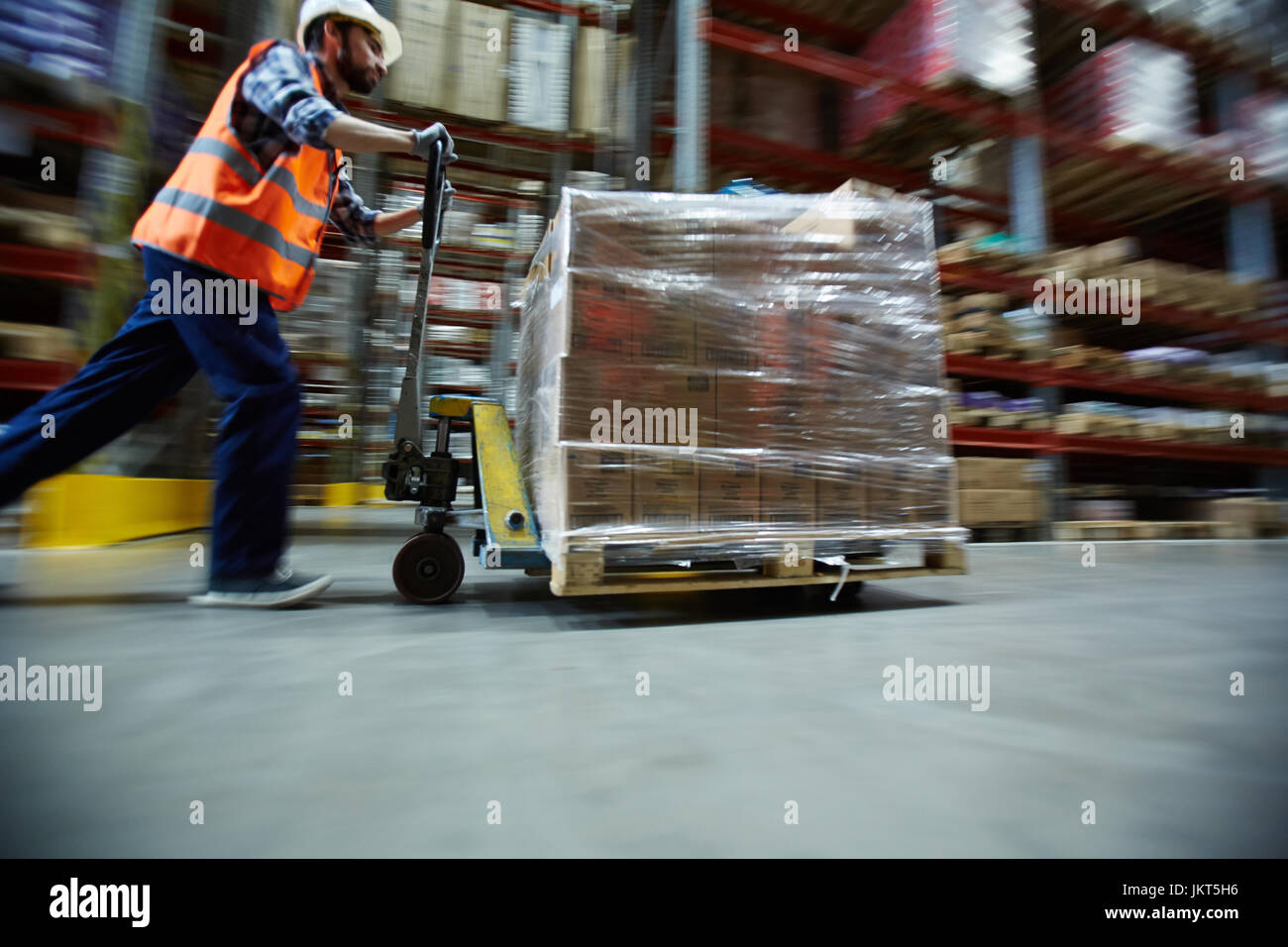 Sfocata colpo di movimento di magazzino Lavoratore che indossa hardhat e giubbotti catarifrangenti spingendo il carrello in movimento con scatole lungo isola tra alte scaffalature Foto Stock