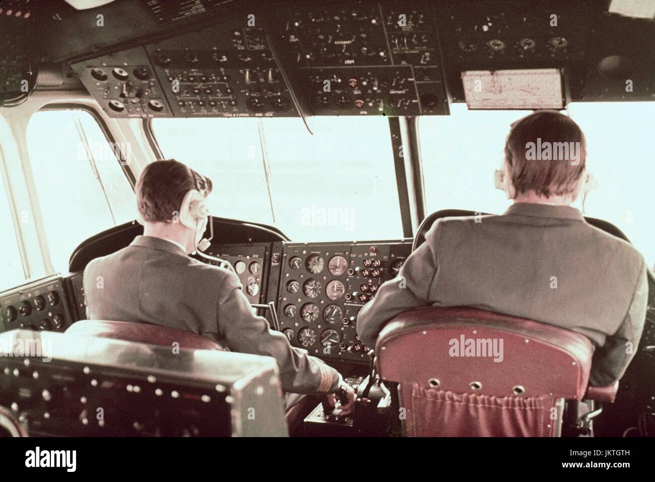 Due formalmente vestito piloti indossare cuffie di comunicazioni sedersi nella cabina di pilotaggio di un aereo passeggeri, con un pilota che si allunga verso il comando della valvola a farfalla, in aria, 1970. Foto Stock