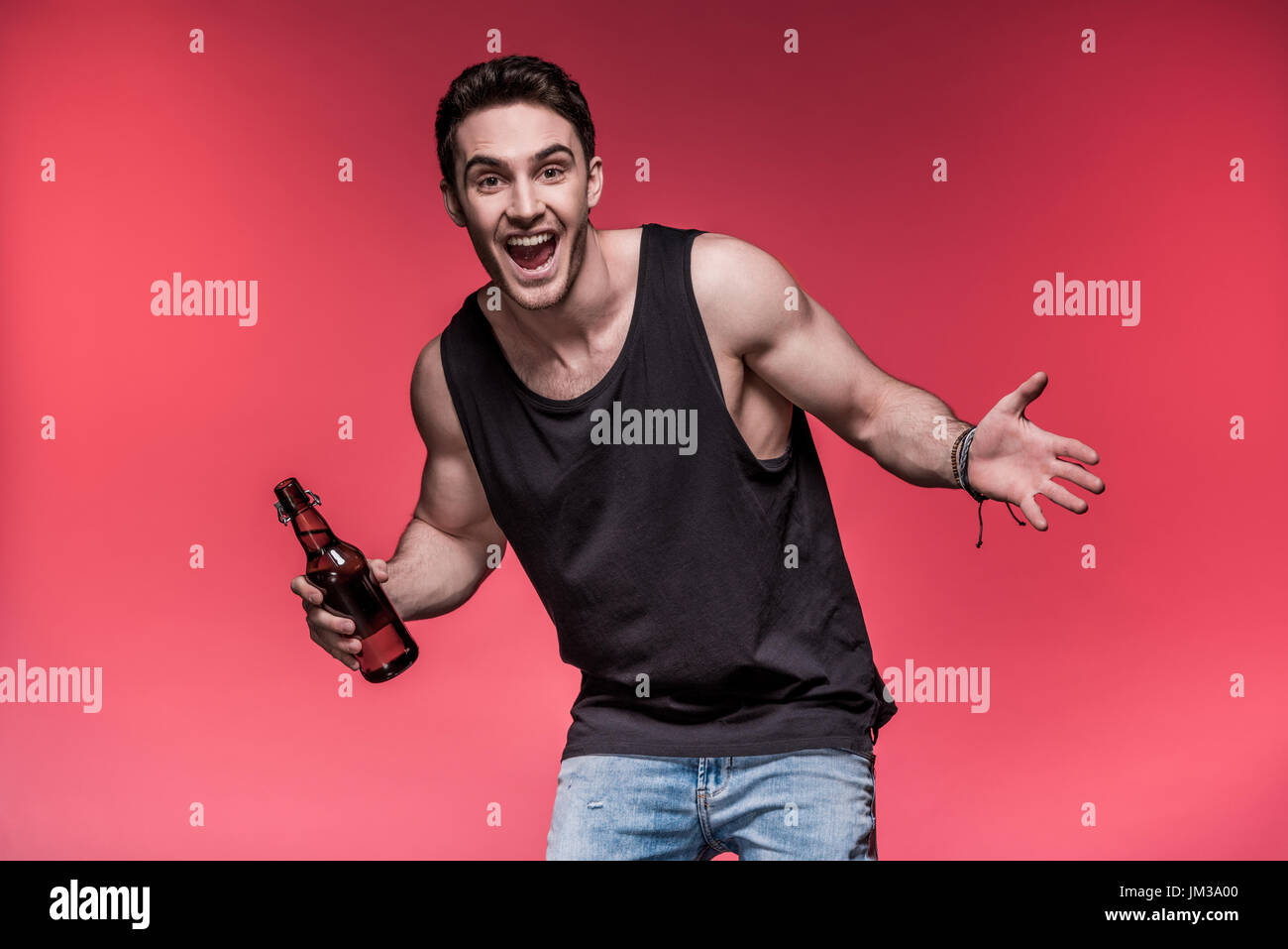 Bel giovane gesticolando con bottiglia di birra e sorridente in telecamera Foto Stock