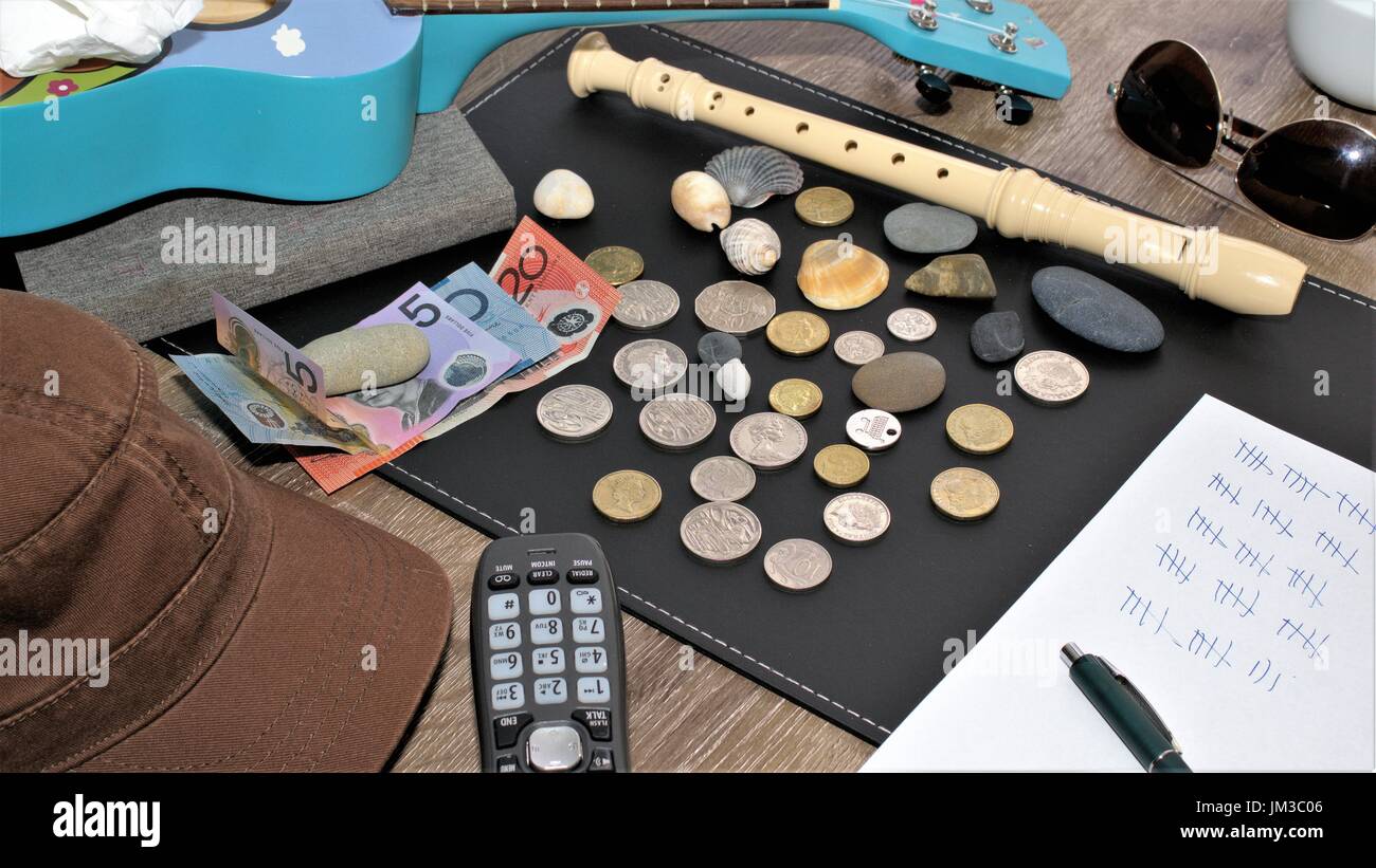 Dollaro australiano note e centesimi di dollaro australiano, carta penna scrittura, hat, cappuccio, ukulele, scrunched il tessuto, flauto, occhiali da sole, telefono cordless su un tavolo Foto Stock