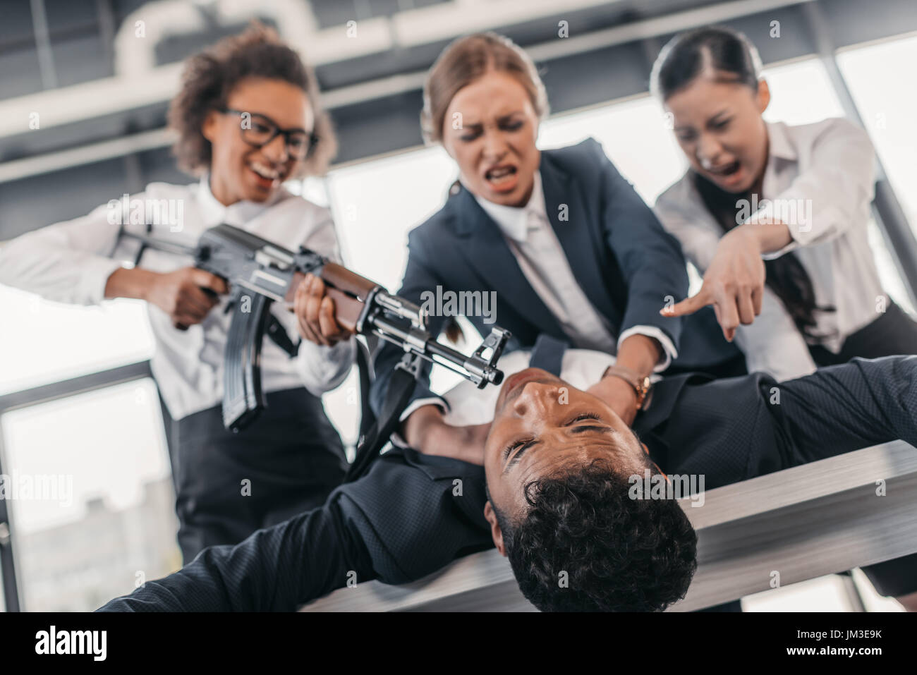Tre giovani imprenditrici arrabbiato con un fucile punire l uomo d affari sdraiato sul tavolo, riunioni di team aziendali concetto Foto Stock
