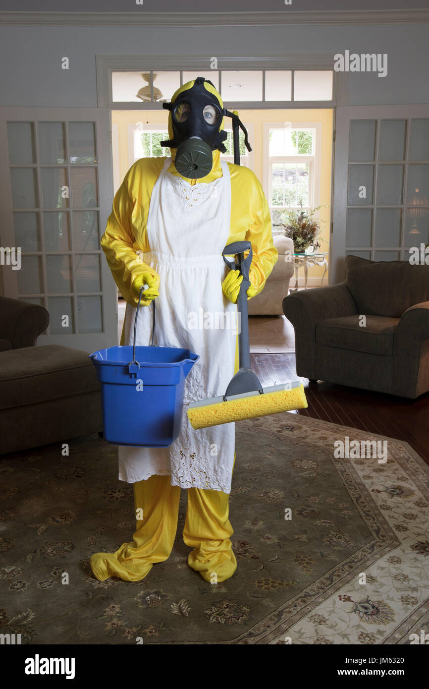 Donna matura in giallo Haz Mat suir e maschera a gas in piedi in salotto con benna blu e spugna mop. Foto Stock