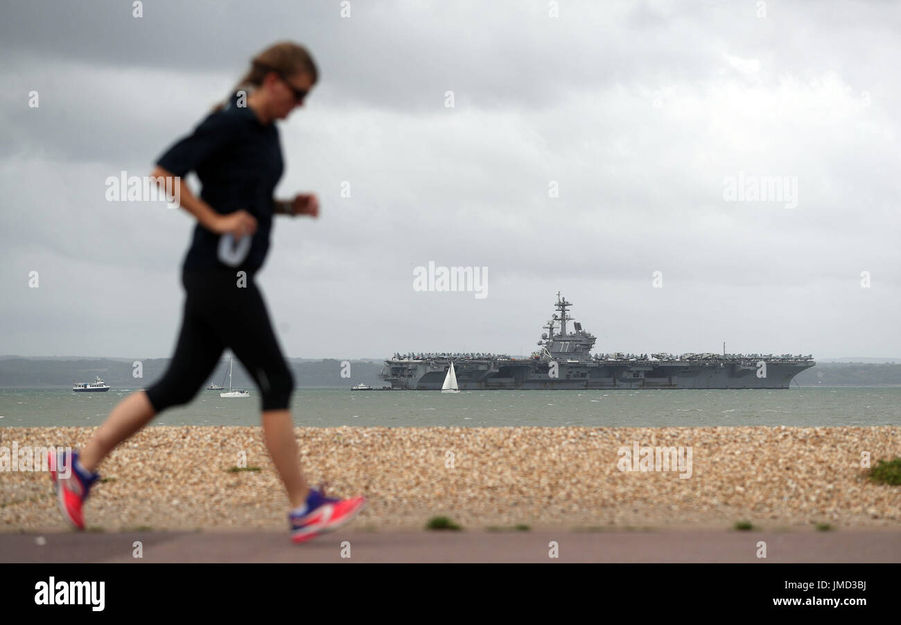 Una persona che corre lungo la spiaggia di ciottoli di Stokes Bay, Hampshire, come la US Navy Nimitz-class portaerei USS George H.W. Boccola fissa ancorata al largo prima di esso partecipa n esercizio guerriero sassone 2017 nel Nord dell'Oceano Atlantico. Foto Stock