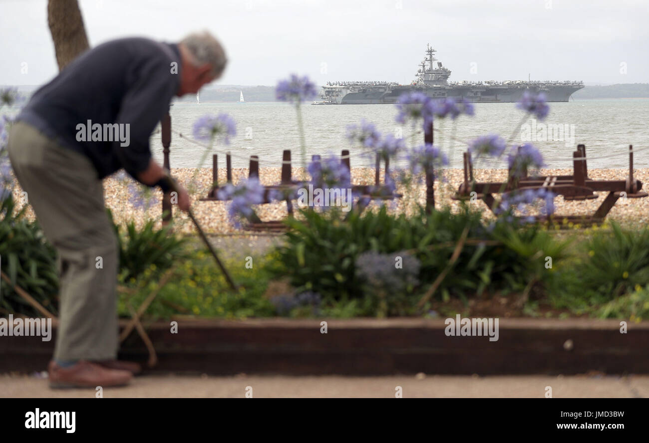 Una persona tende a un letto di fiori sulla spiaggia di ciottoli di Stokes Bay, Hampshire, come la US Navy Nimitz-class portaerei USS George H.W. Boccola fissa ancorata al largo prima di esso partecipa n esercizio guerriero sassone 2017 nel Nord dell'Oceano Atlantico. Foto Stock